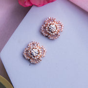 A pair of rose gold-colored sterling silver stud earrings with a floral bloom motif, placed on a white surface with a pink flower and a pale background.